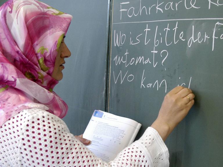 Eine Lehrerin mit Kopftuch steht an einer Tafel und schreibt deutsche Sätze auf.