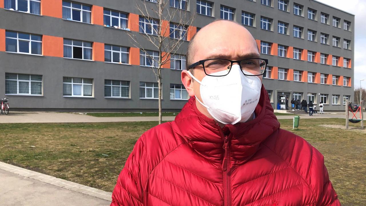 Philipp Eysel trägt eine rote Jacke und einen Mund-Nasen-Schutz. Er hat eine Brille auf. Im Hintergrund sieht man ein breites, schlichtes Gebäude, die Fassade ist in grau und rot gehalten.
