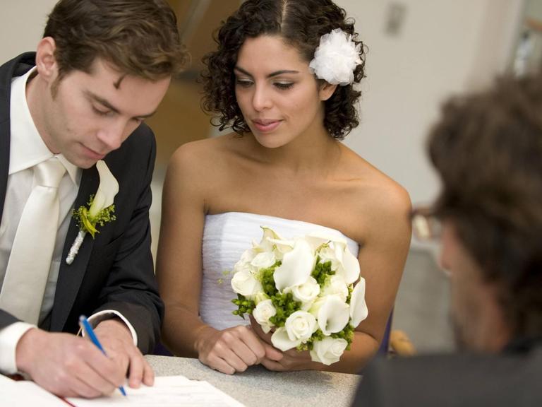 Standesamtliche Hochzeit: Der Bräutigam unterschreibt Heiratsurkunde, daneben die Braut mit Blumengebinde