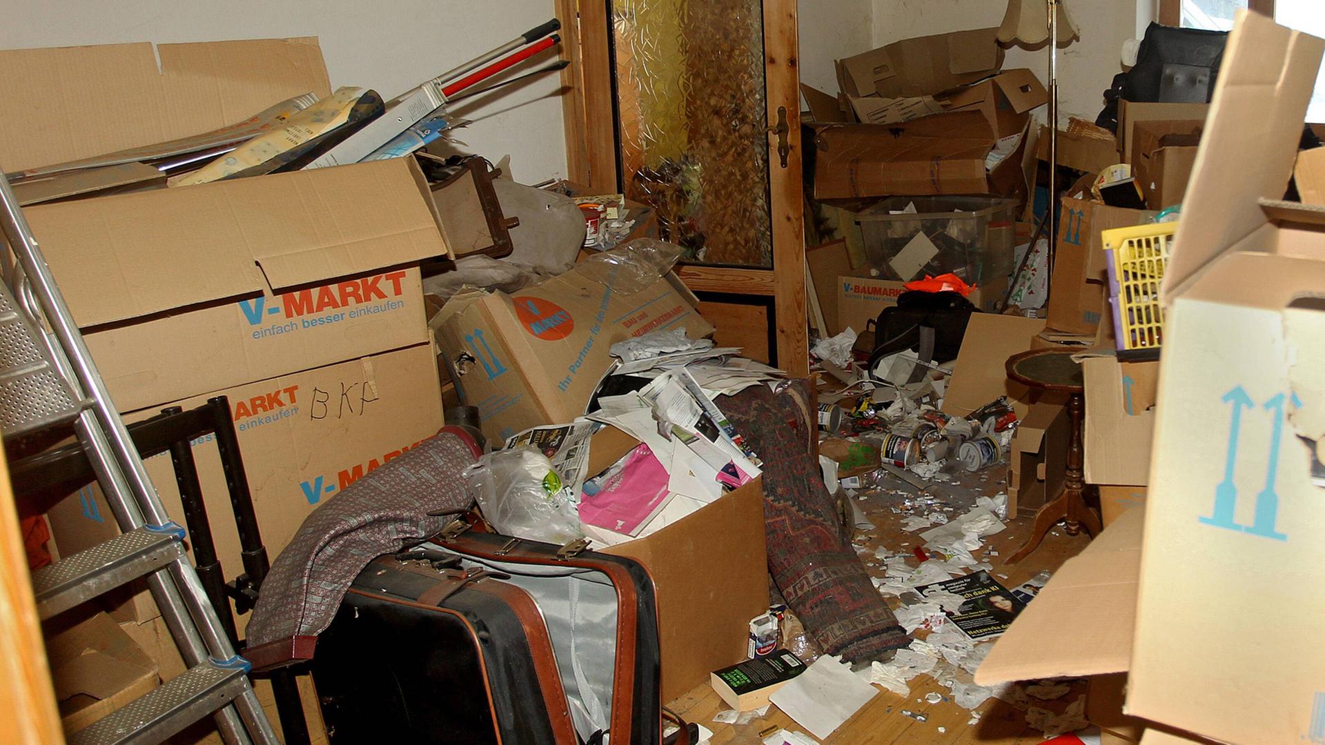Eine vermüllte Wohnung: Kisten, eine Leiter, ein alter Koffer und viele weitere Gegenstände sind auf dem Boden zu sehen.
