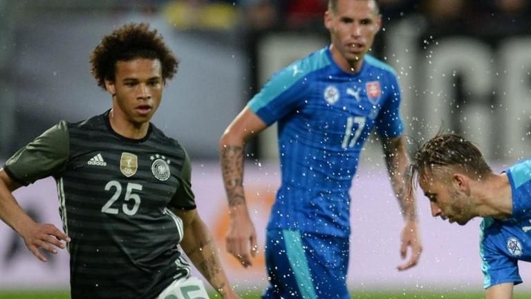 Sie sehen den Fußball-Spieler Leroy Sané, er ist links auf dem Bild. Er kämpft gegen einen Spieler aus dem Land Slowakei. Sie sehen den Fußball-Spieler Leroy Sané, er ist links auf dem Bild. Er kämpft gegen einen Spieler aus dem Land Slowakei.