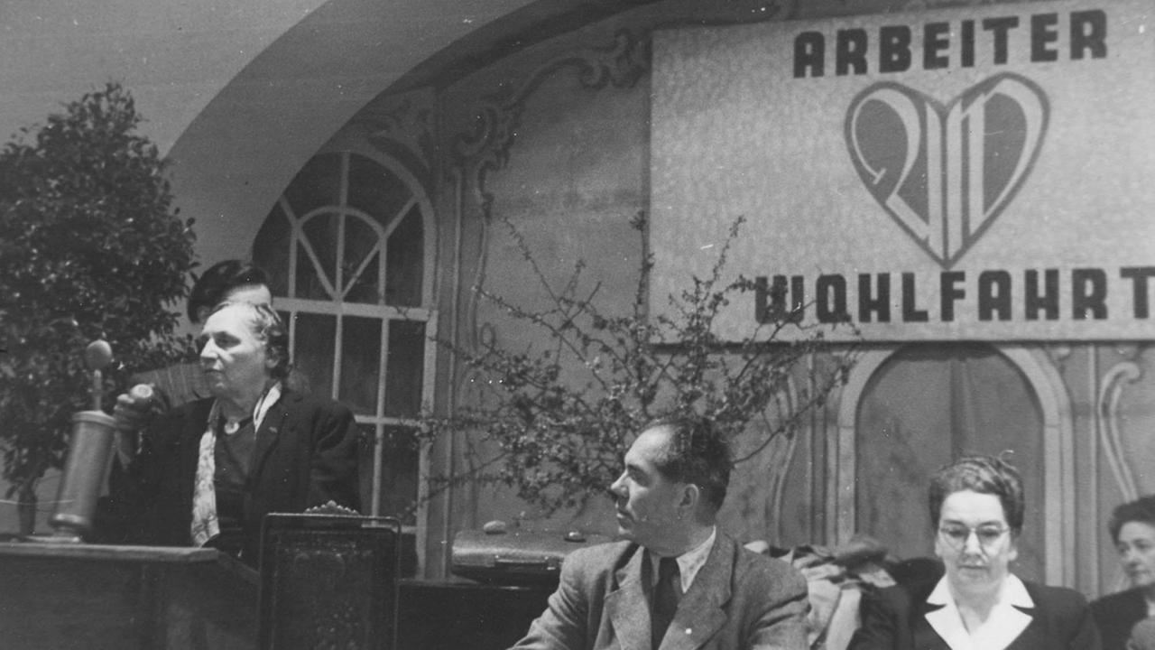 AWO-Gründerin Marie Juchacz (l), Franz Neumann und Ida Wolff Ende der 40er-Jahre auf einer Veranstaltung der Arbeiterwohlfahrt. Eine Frau steht an einem Rednerpult, daneben sitzen ein Mann und eine Frau.