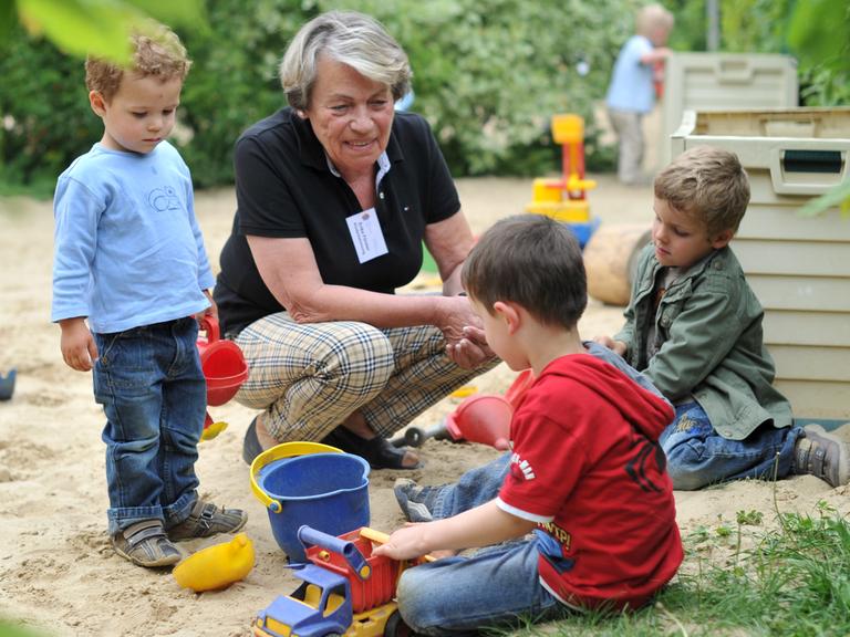 Eine Seniorin spielt im Garten des Mehrgenerationenhauses mit mehreren Kindern in einem Sandkasten.