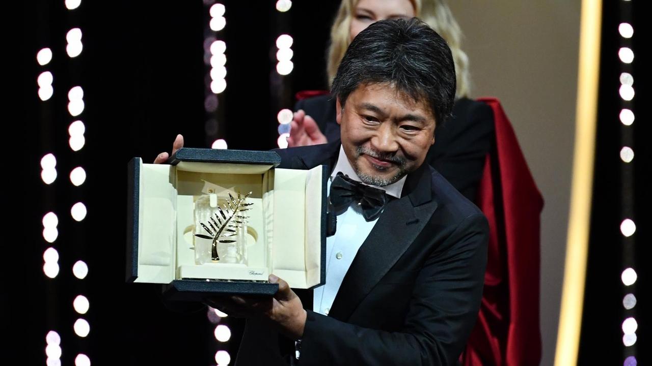 Auf dem Bild sieht man den Filme-Macher Kore-eda Hirokazu. Er kommt aus dem Land Japan. Er hält einen Preis in der Hand. Den hat er für seinen Film gewonnen. 