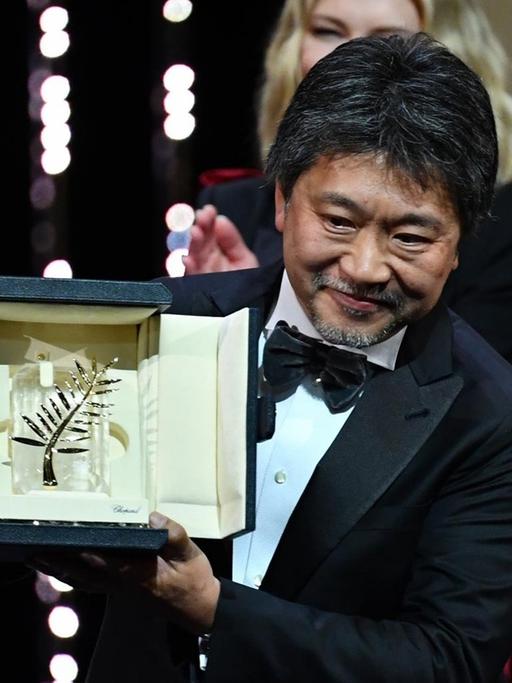 Auf dem Bild sieht man den Filme-Macher Kore-eda Hirokazu. Er kommt aus dem Land Japan. Er hält einen Preis in der Hand. Den hat er für seinen Film gewonnen. 