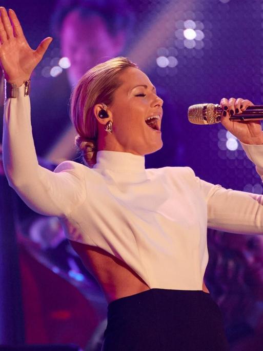 Die Sängerin Helene Fischer agiert beim steht mit Mikrofon und in weißem Oberteil auf einer Bühne und singt.