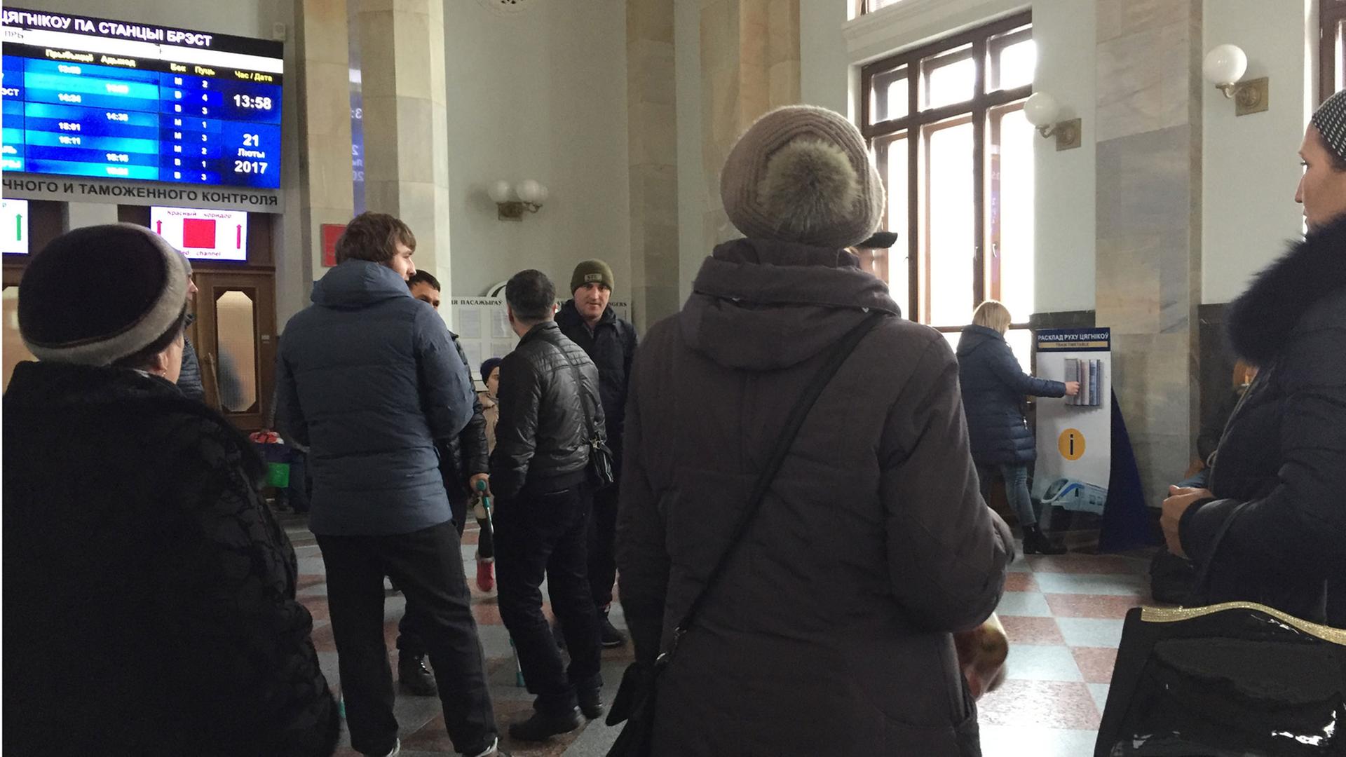 Das Bild zeigt wartende Tschetschenen am Bahnhof von Brest von hinten. Sie schauen auf eine Anzeigetafel im Bahnhof. Von dort aus versuchen viele Menschen, über Polen in die Europäische Union zu gelangen.