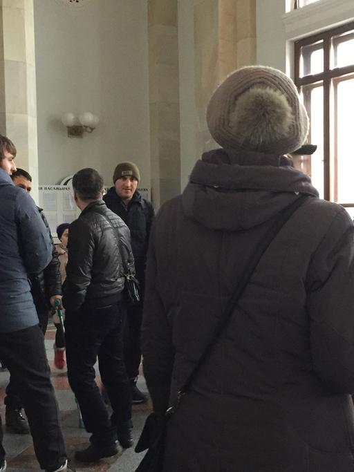 Das Bild zeigt wartende Tschetschenen am Bahnhof von Brest von hinten. Sie schauen auf eine Anzeigetafel im Bahnhof. Von dort aus versuchen viele Menschen, über Polen in die Europäische Union zu gelangen.