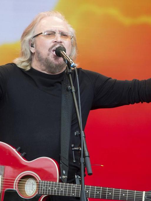 Barry Gibb von den Bee Gees, mit langen grauen Haaren. Er hat eine rote Gitarre um den Hals und breitet die Arme weit aus. Der Hintergrund ist rot-orange gefärbt und erinnert an einen Sonnenaufgang.