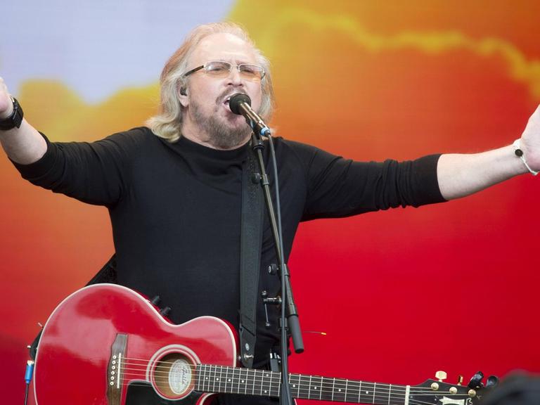 Barry Gibb von den Bee Gees, mit langen grauen Haaren. Er hat eine rote Gitarre um den Hals und breitet die Arme weit aus. Der Hintergrund ist rot-orange gefärbt und erinnert an einen Sonnenaufgang.