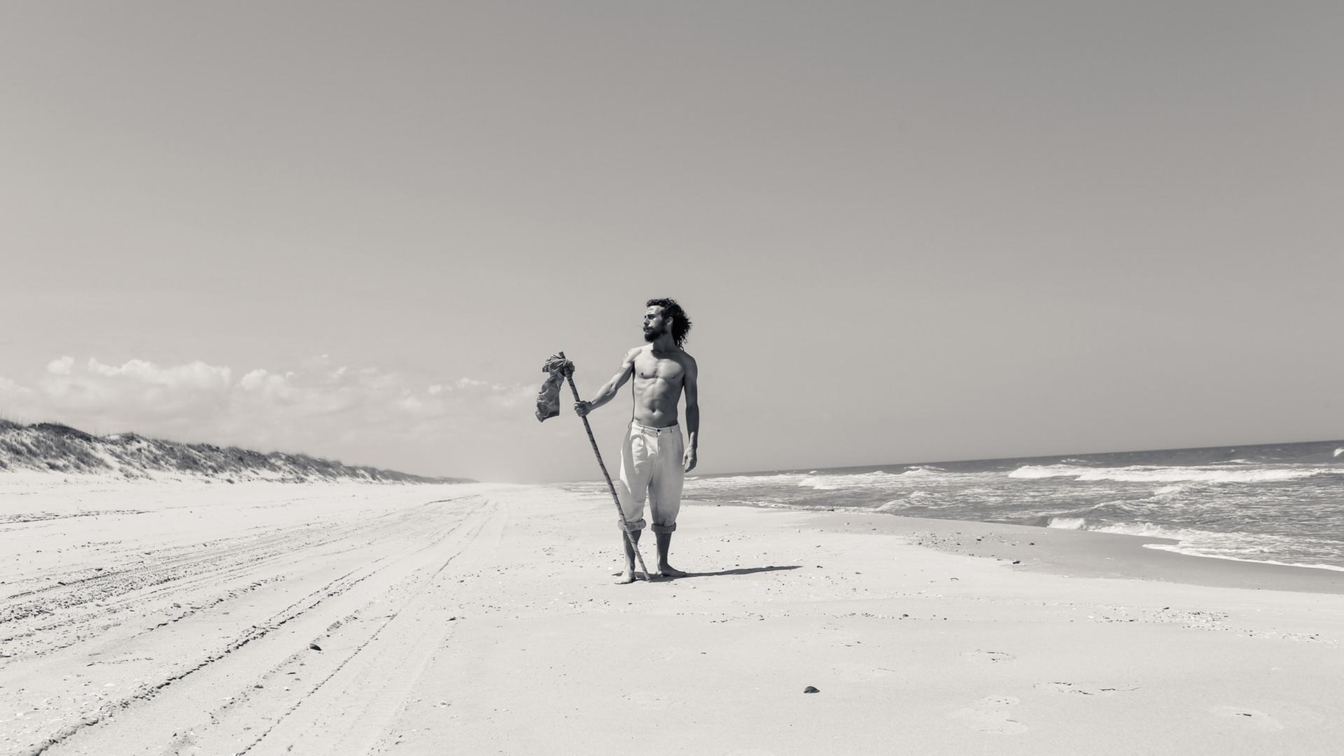 Ein Mann mit langen Haaren und Bart steht, nur mit einer kurzen Hose bekleidet, an einem Meeresstand. Er hält einen Stock in der Hand und schaut in die Ferne.