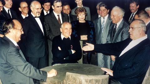 Bundeskanzler Helmut Kohl (r), der sowjetische Staatspräsident Michail Gorbatschow (M) und Bundesaußenminister Hans-Dietrich Genscher (l) unterhalten sich am 15.07.1990 in entspannter Atmosphäre an einem rustikalen Arbeitstisch in der freien russischen Natur, während die anderen Gäste unter anderem Raissa Gorbatschowa (M hinten) und Eduard Schewardnadse amüsiert die Szene betrachten.