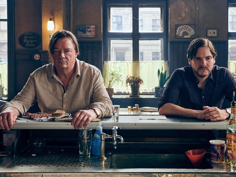 Peter Kurth und Daniel Brühl in einer Szene aus dem Film Nebenan. Sie sitzen nebeneinander an einer Theke in einer Bar.