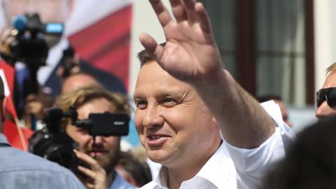 Nahaufnahme von Duda, der lächelnd winkt. Um ihn herum ein Kameramann und weitere Personen. Ganz hinten unscharf ein Wahlkampfplakat Dudas.