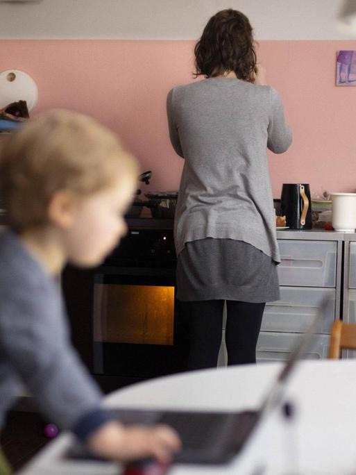 Eine Frau steht mit dem Rücken zum Betrachter in einer modernen Küche am Herd. Im Vordergrund ist in der Unschäre ein kleiner Junge an einem Laptop zu sehen.