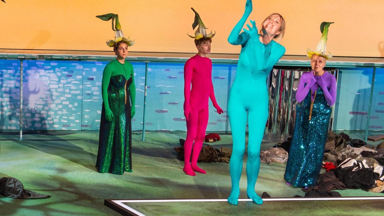 Drei Frauen und ein Mann stehen auf der Theaterbühne. Sie tragen einfarbige Gewänder in den Farben Grün, Pink, Türkis und Lila, drei Personen tragen Hütchen auf dem Kopf die aussehen wie umgedrehte große Papierblumen mit Stiel.