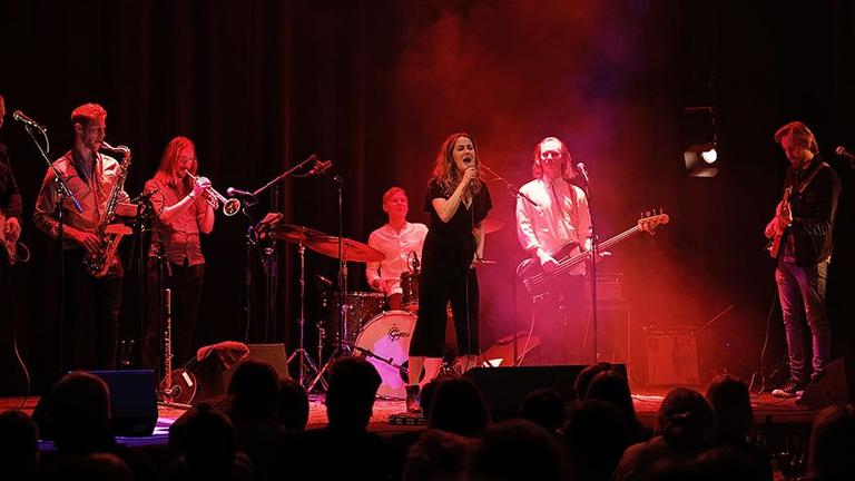 Eine Sängerin steht mit ihrer Band, die aus sechs Männern besteht, auf einer rot erleuchteten Bühne.