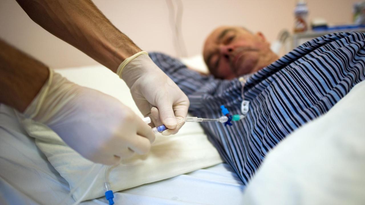 Zu sehen sind die Hände des Pflegers mit Gummihandschuhen, sie befestigen eine Kanüle, dahinter der im Bett liegende Patient. 
