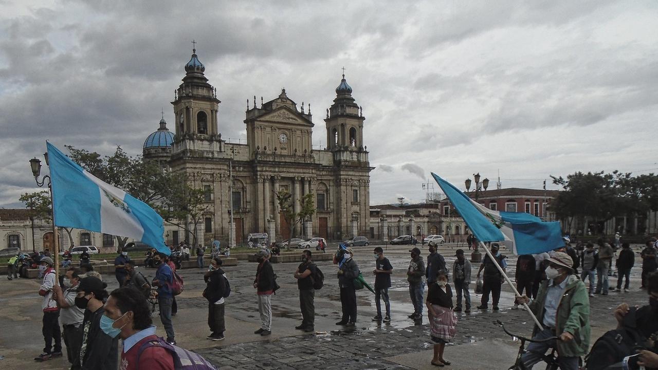 Demonstrierende stehen auf dem Platz vor der Kathedrale in Guatemala-Stadt, zwei von ihnen tragen blau-weiße Fahnen, alle tragen Mund-Nase-Masken und halten Abstand voneinander, um sich während der weltweiten Covid-19-Pandemie vor Ansteckung zu schützen, der Himmel ist wolkenverhangen.