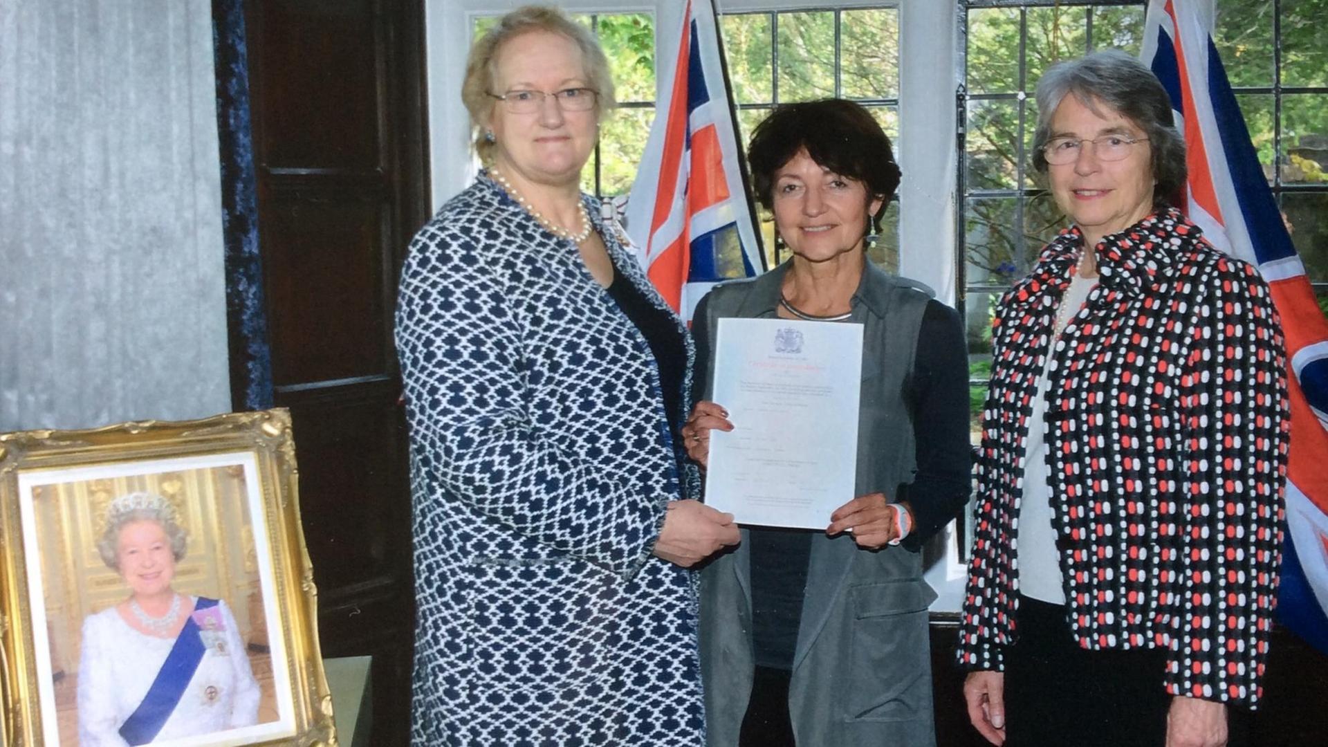 Ruth Rach bei ihrer Einbürgerung im südenglischen Städtchen Lewes. Links neben ihr steht die Vertreterin der Königin, rechts eine Verwaltungsbeamtin. Links unten ein Bild Königin Elizabeth II.