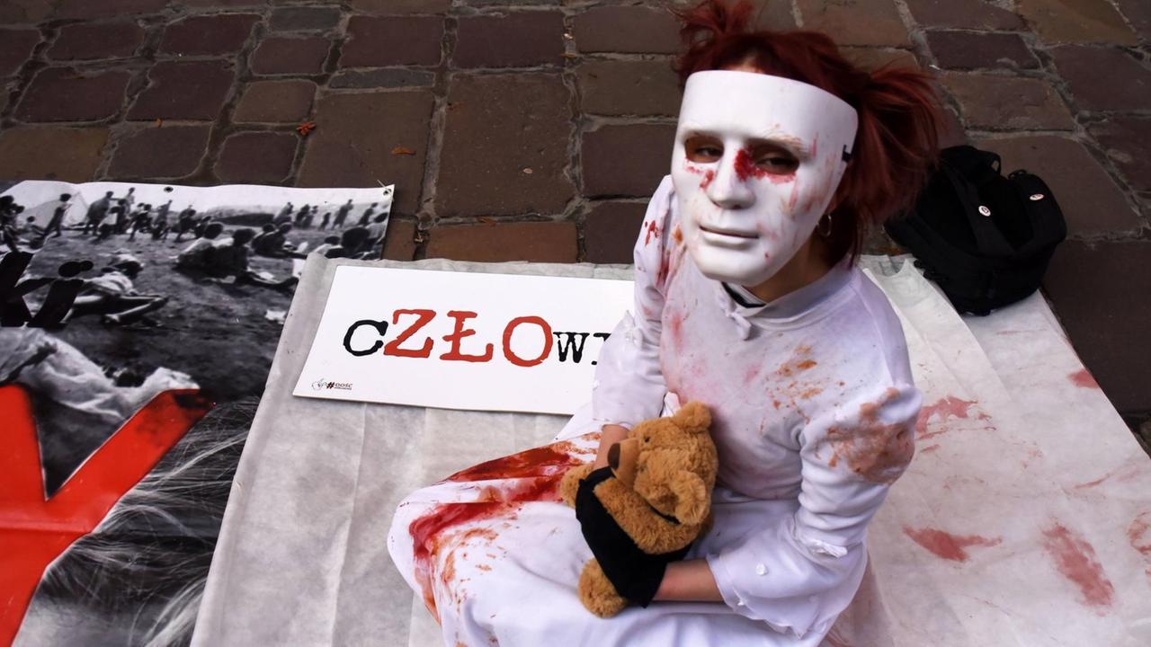 Darstellerin sitzt mit Maske, blutverschmiertem Kittel und Teddybär auf dem Asphalt vor dem Bischofspalast in Krakau am 22. November
