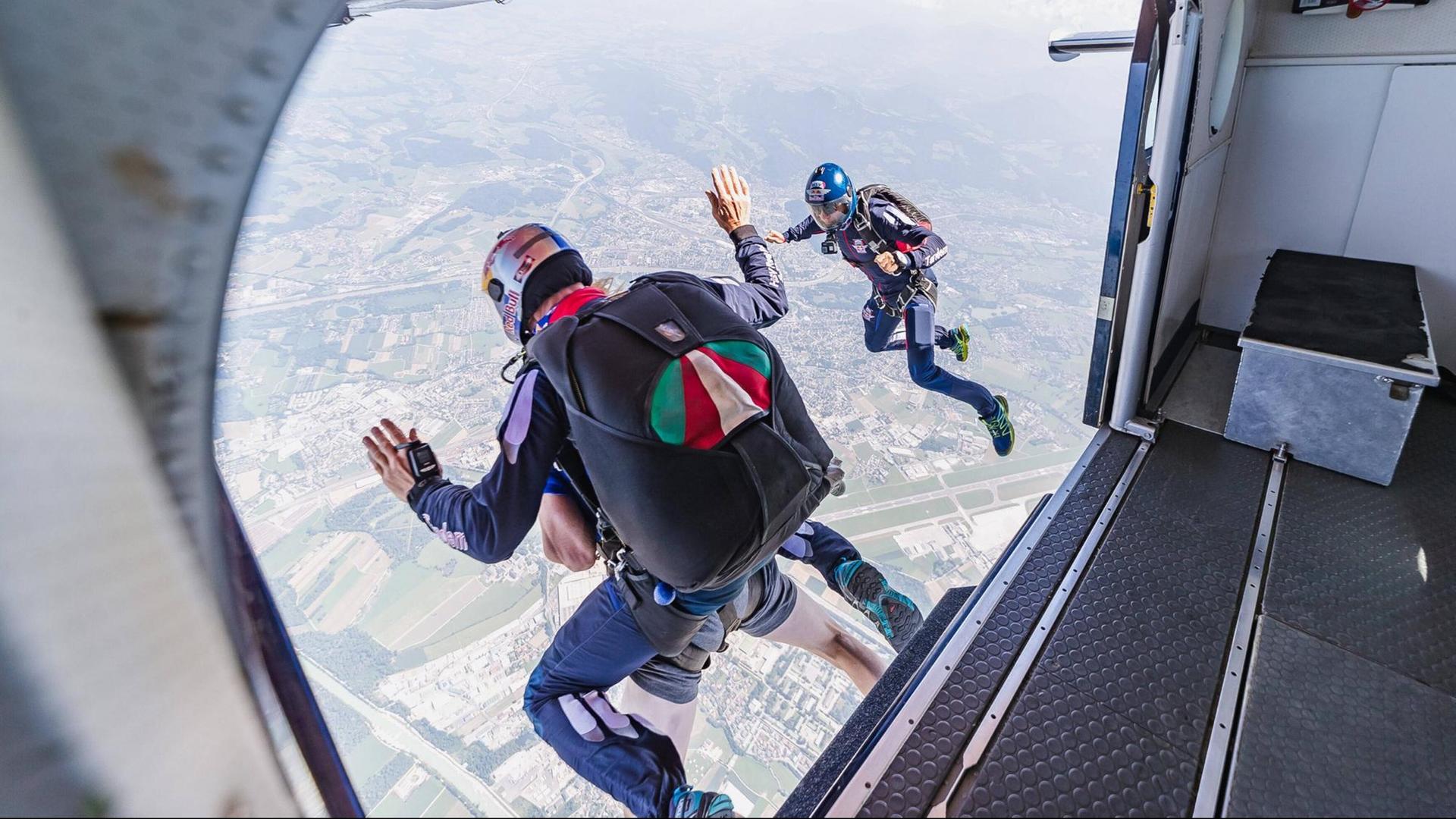 Drei Personen mit Fallschirmen springen aus einem Flugzeug. Unten sieht man eine bebaute Landschaft.