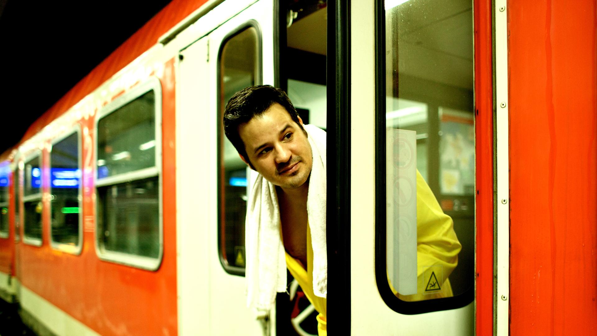 René El Khazraje alias MC Rene zelebriert das Leben im Zug. Auf dem Bild schaut er im Bademantel und Handtuch aus der Tür eines Regionalexpresses der Bahn.
