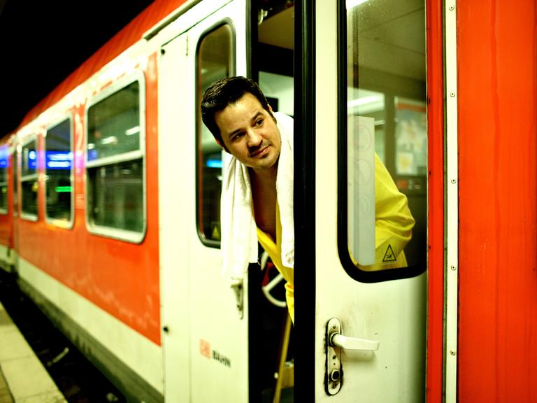 René El Khazraje alias MC Rene zelebriert das Leben im Zug. Auf dem Bild schaut er im Bademantel und Handtuch aus der Tür eines Regionalexpresses der Bahn.