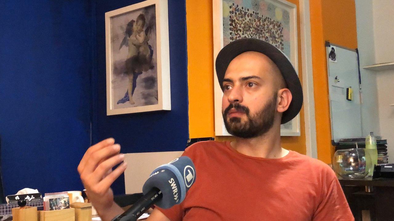 Maziyar Khavajian sitzt in seiner Wohnung mit Hut, schwarzem Vollbart und dunkelrotem T-Shirt vor dem Mikrofon und gestikuliert mit der rechten Hand.