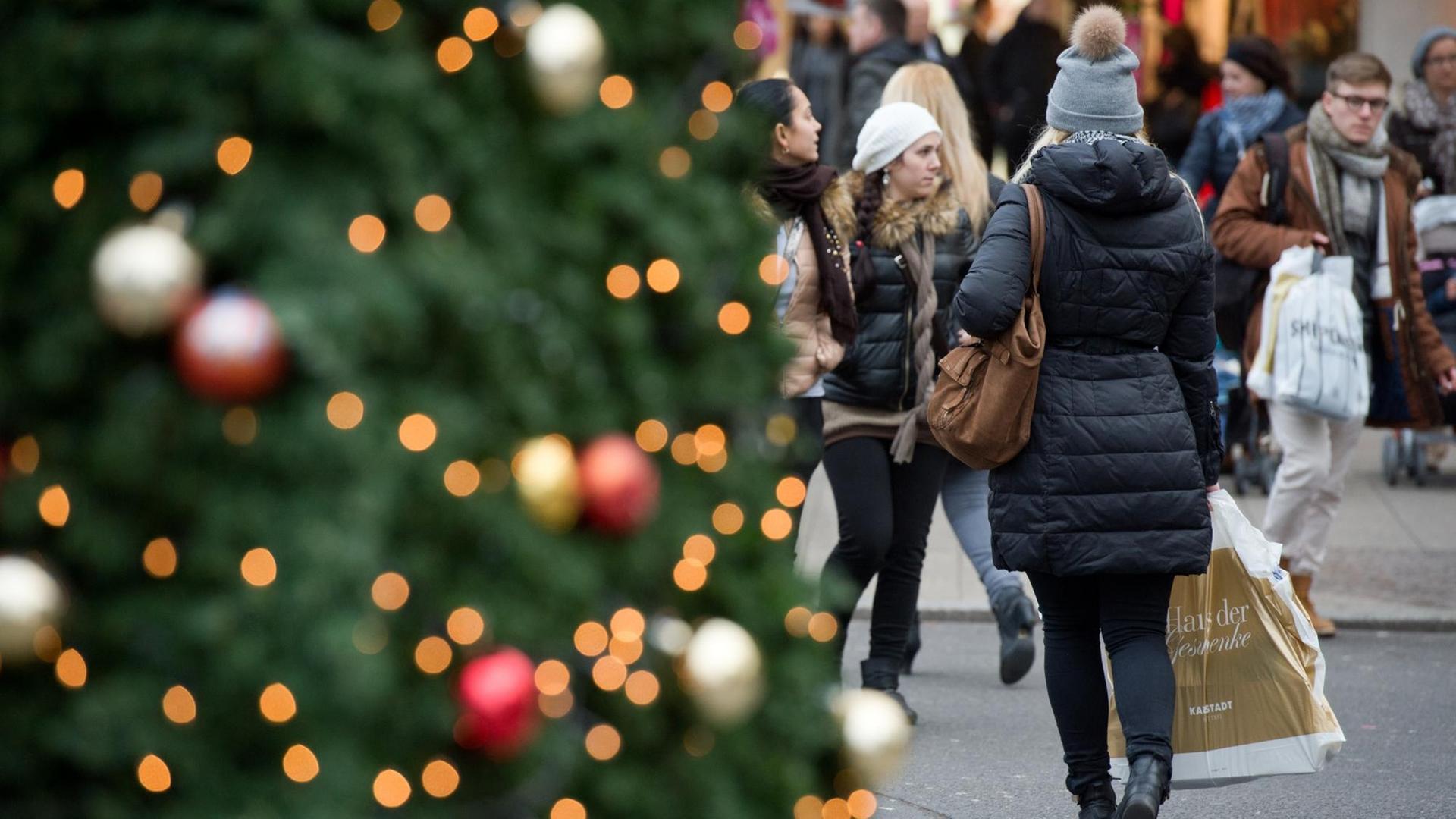 Links im Vordergrund ein Weihnachtsbaum neben dem eine Frau mit einer Einkaufstüte entlanggeht.