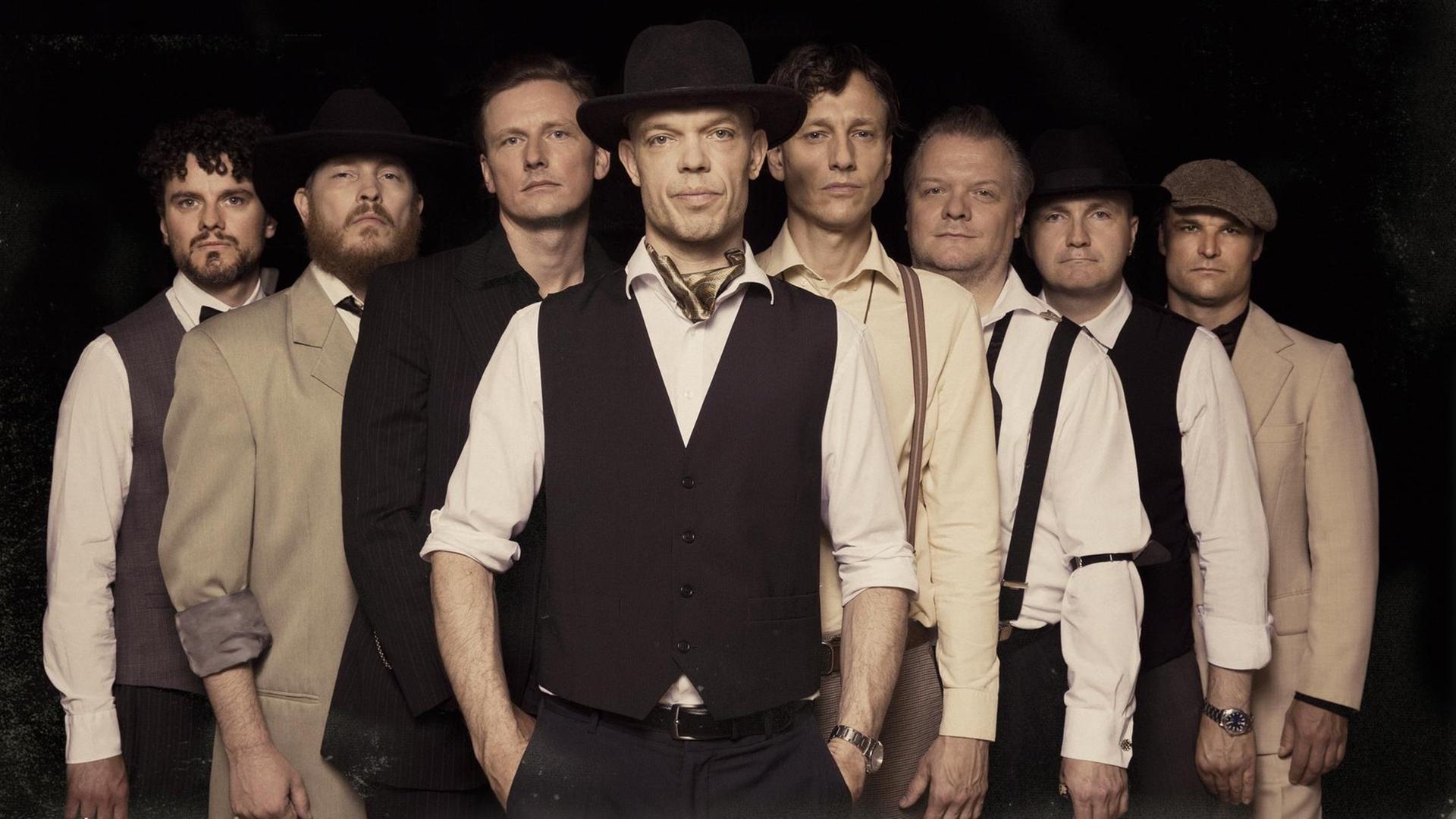 Ein Band aus acht Musikern steht V-förmig, an der Spitze der dänische Gitarrist und Sänger Thorbjørn Risager mit weißem Hemd, Hut und schwarzer Weste.