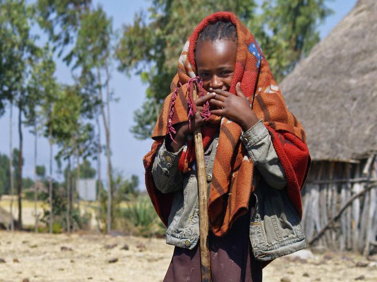 Mädchen mit Decke um den Kopf und Holzstock in der Hand vor einer Hütte in Uganda