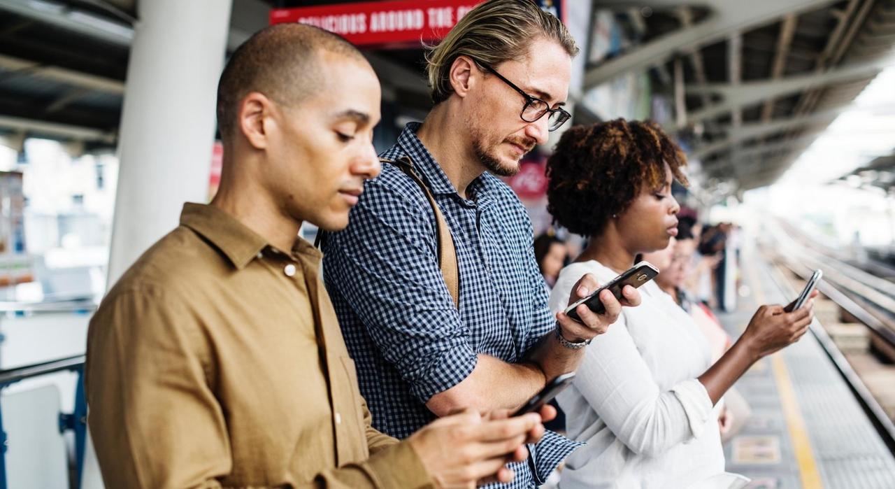 Drei Menschen stehen nebeneinander auf einem Bahnsteig und gucken auf ihre Smartphones