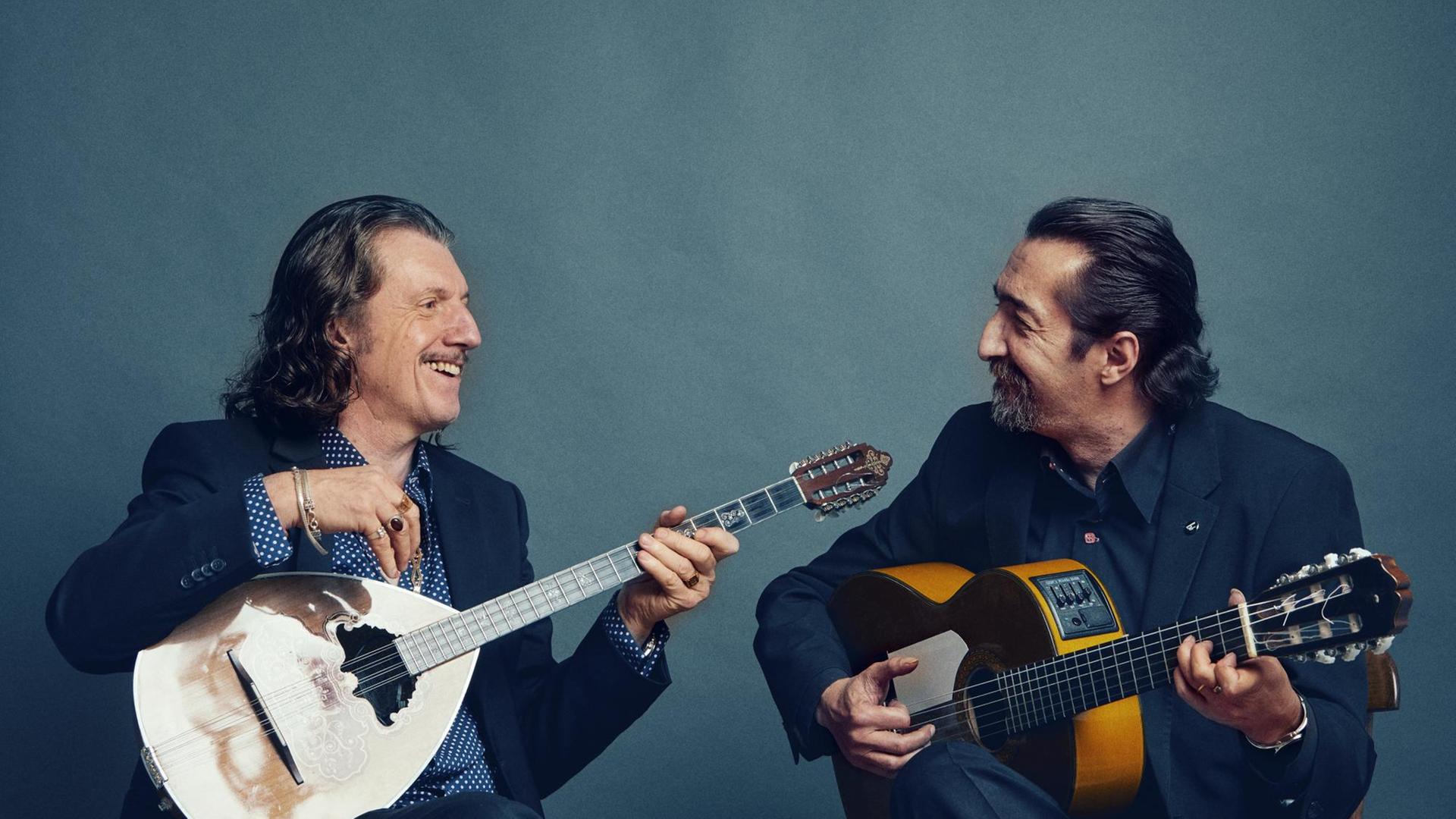 Zwei Männer sitzen auf Hockern. Der linke Mann hält eine Buzuk, der rechte Mann eine Gitarre in der Hand.