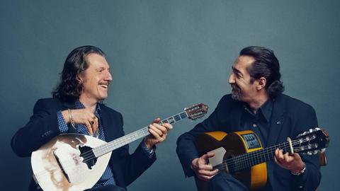 Zwei Männer sitzen auf Hockern. Der linke Mann hält eine Buzuk, der rechte Mann eine Gitarre in der Hand.