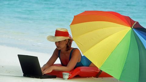 Eine Frau liegt unter einem bunten Sonnenschirm an einem Sandstrand und arbeitet an ihrem Laptop.