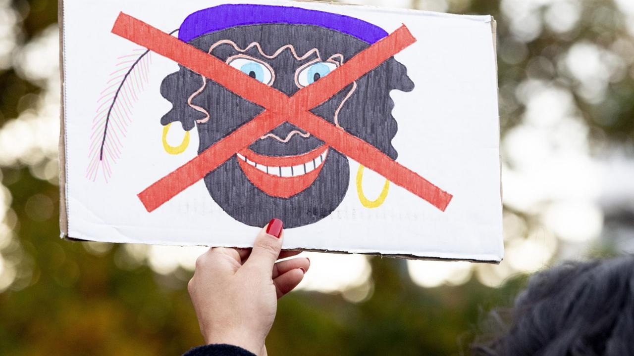 Eine Hand mit roten Fingernägeln hält ein Protestplakat, auf dem ein gezeichnetes Bild eines schwarz geschminkten "Zwarten Piet" mit goldenen Kreolen und violetter Mütze rot durchgestrichen ist.