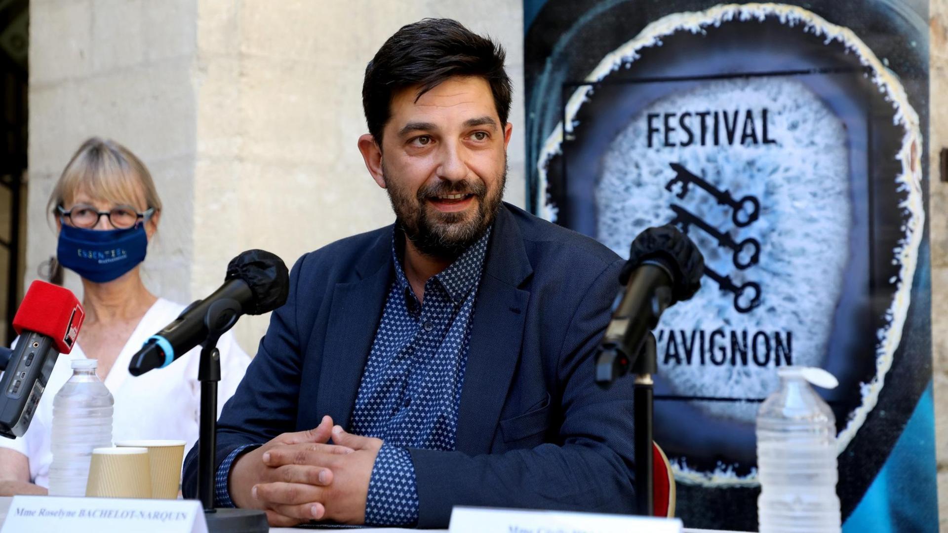 Festivalleiter Tiago Rodrigues sitzt im Sommeranzug vor Mikrofonen, hinter sich ein Schild des Theaterfestivals Avignon mit drei Schlüsseln.