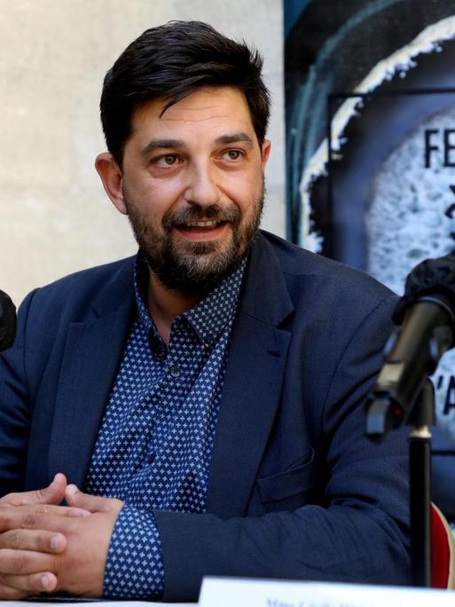 Festivalleiter Tiago Rodrigues sitzt im Sommeranzug vor Mikrofonen, hinter sich ein Schild des Theaterfestivals Avignon mit drei Schlüsseln.