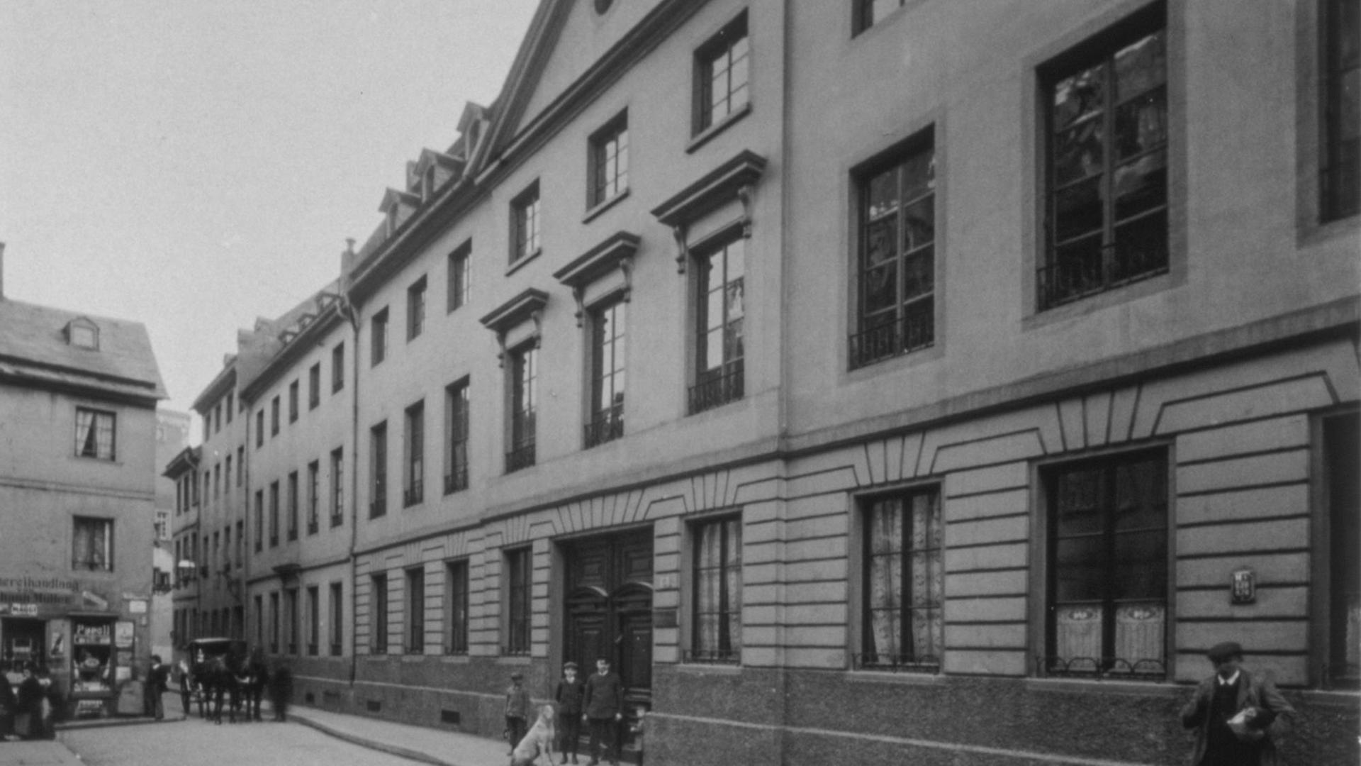 Auf dem s/w Bild ist eine belebte Straßenszene des Jahres 1919 zu sehen: Eine lange Häuserfront mit vielen Fenstern, im Erdgeschoss Stuck an der Fassade. Im Hintergrund Menschen und eine Kutsche.