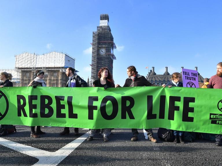 Die Demonstranten stehen nebeneinander auf der Brücke und halten das grüne Banner in den Händen. Dahinter das eingerüstete Parlament mit dem Big Ben.