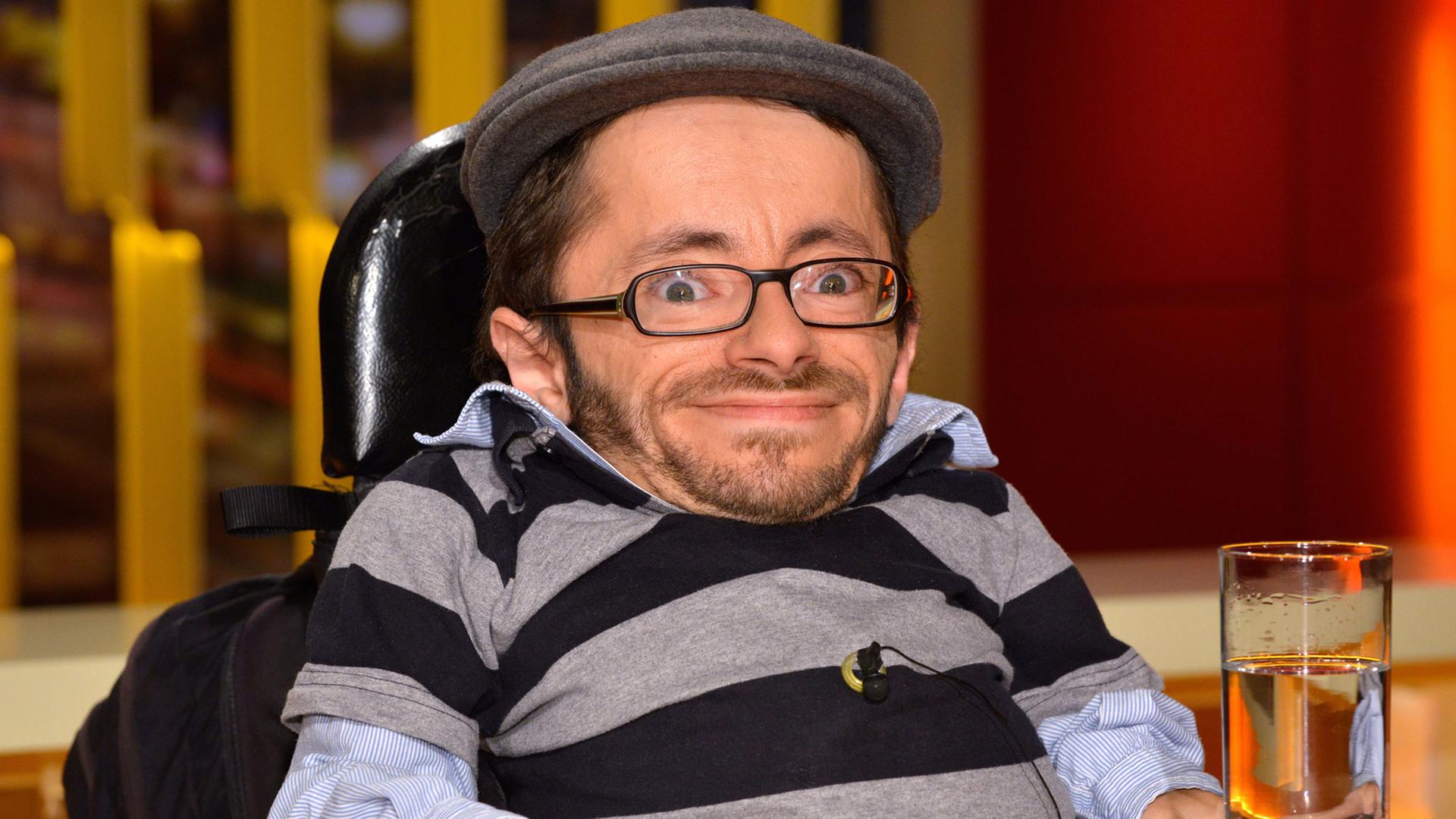 Kleinwüchsiger Mann in einem Rollstuhl mit Brille und Mütze