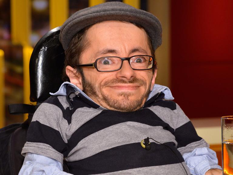 Kleinwüchsiger Mann in einem Rollstuhl mit Brille und Mütze