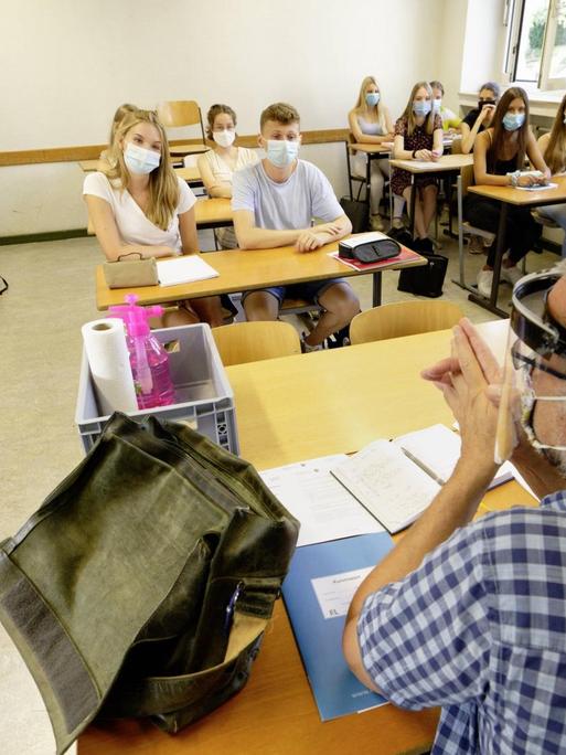 Schülerinnen, Schüler und im Vordergrund ein Lehrer sitzen in einem Klassenraum und tragen Mund-Nasenschutz.