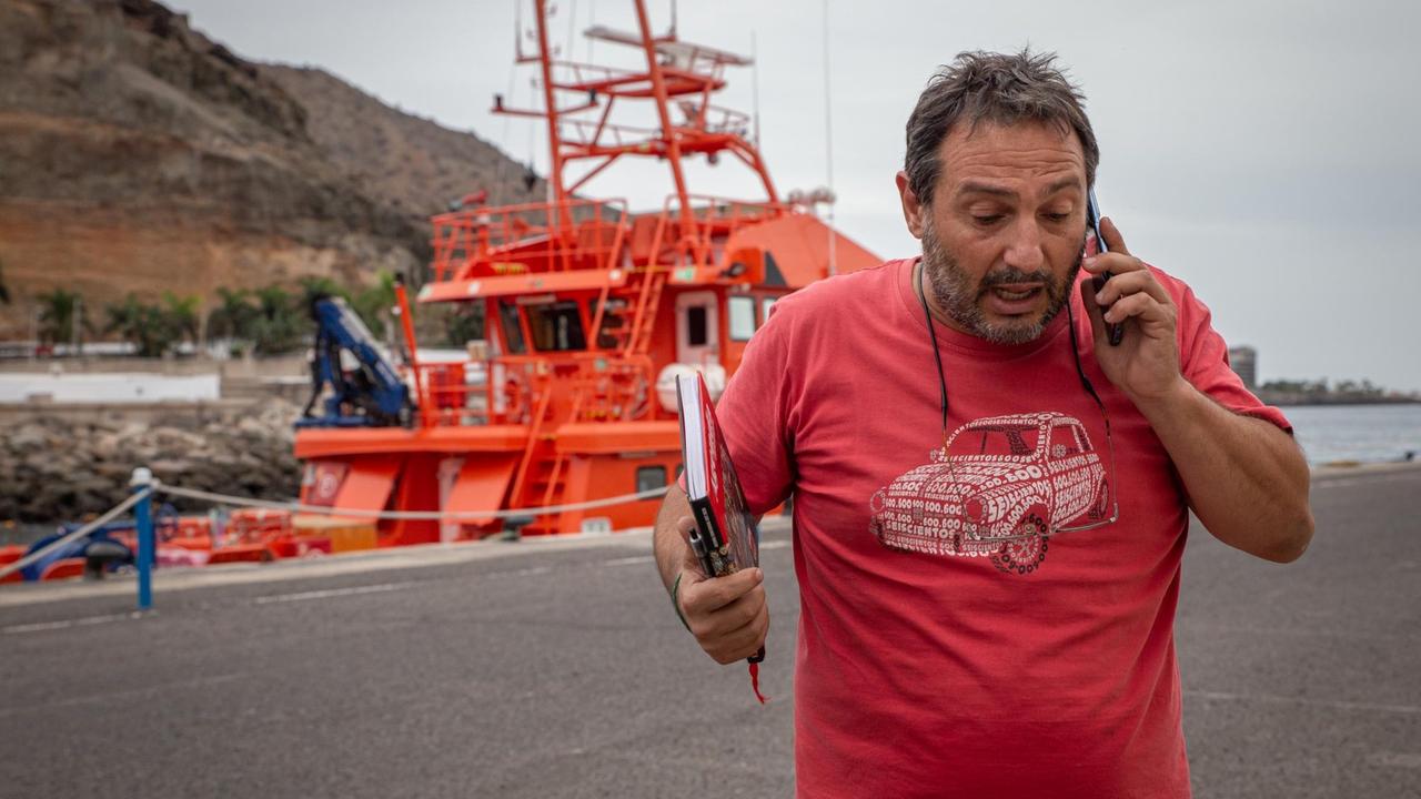 Manuel Capa steht am Hafen und telefoniert gestikulierend. Im Hintergrund sieht man ein knallorange farbenes Rettungsboot.
