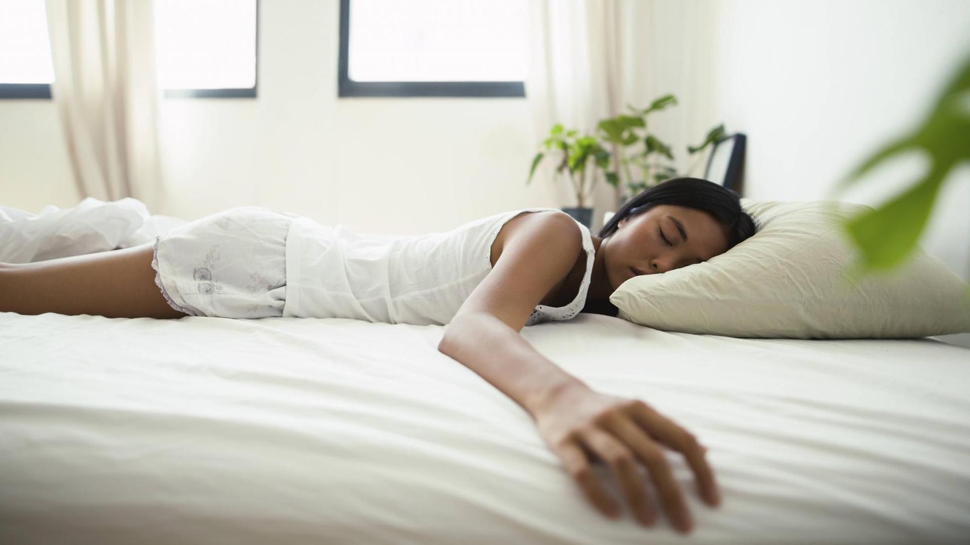 Eine Frau liegt in einem Bett und schläft. Sie liegt dabei auf dem Bauch und streckt einen Arm zur Seite aus.