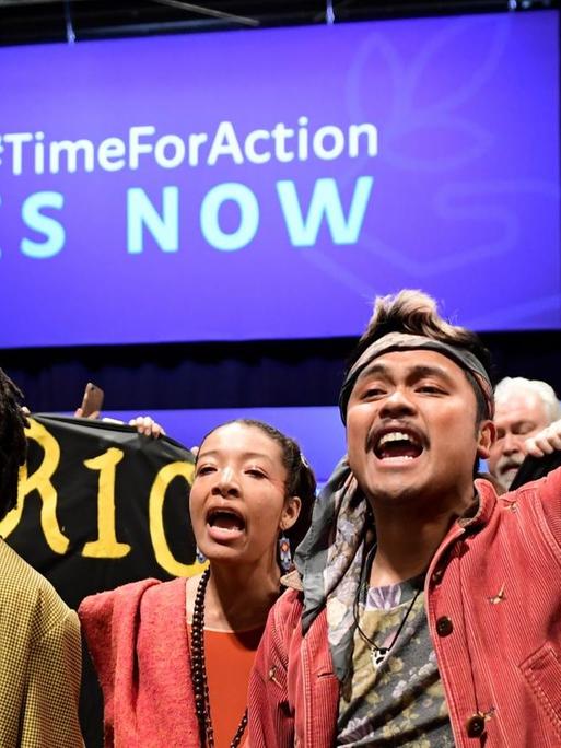 Junge Klimaaktivisten, zum Teil mit hochgestreckten Händen, auf der UN-Klimakonferenz in Madrid. Im Hintergrund ist der Hashtag "TimeForAction, IS NOW" zu sehen.