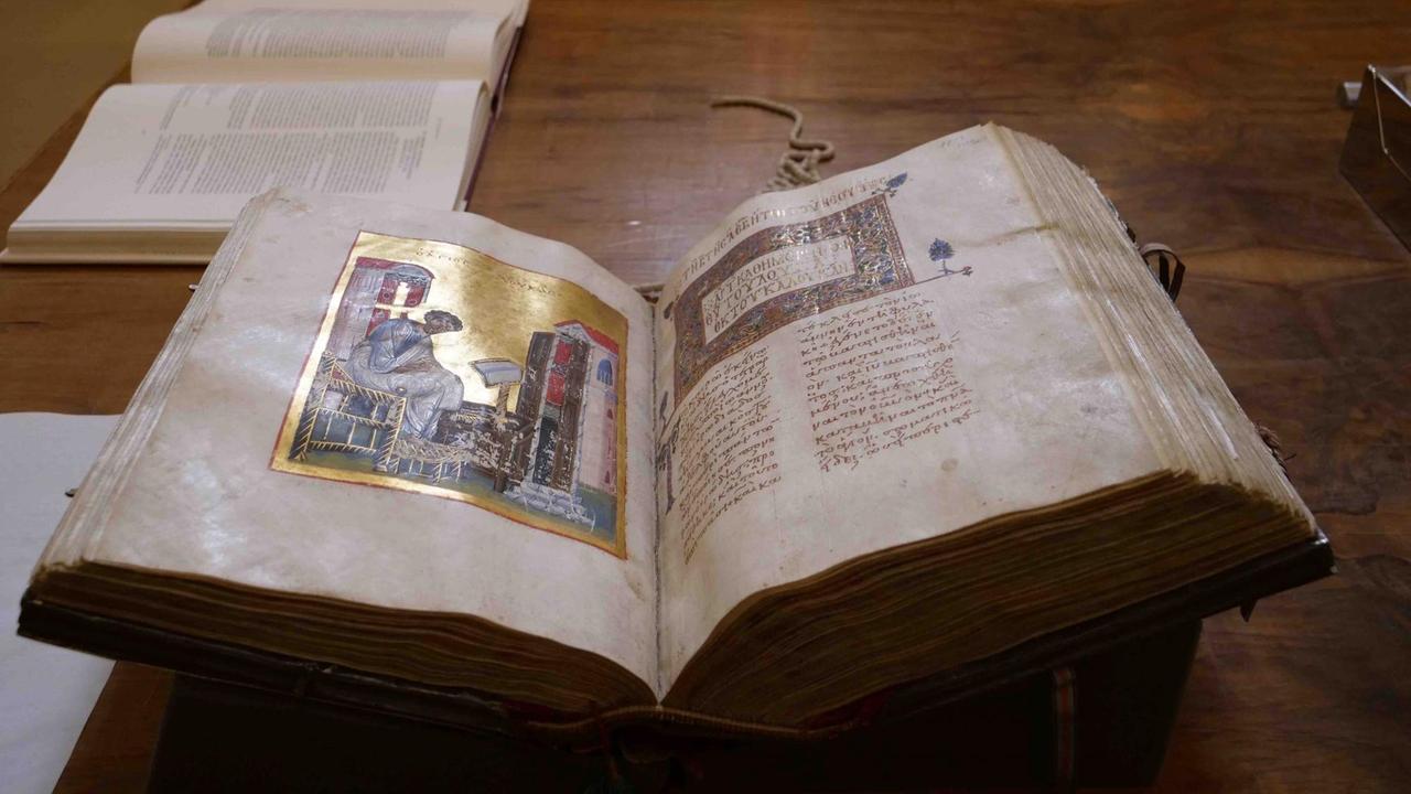 Ein aufgeschlagenes dickes Buch mit leicht angegilbten Seiten zeigt auf der linken Seite eine Illustration mit goldenem Hintergrund, auf der eine Andachtsfigur zu sehen ist.