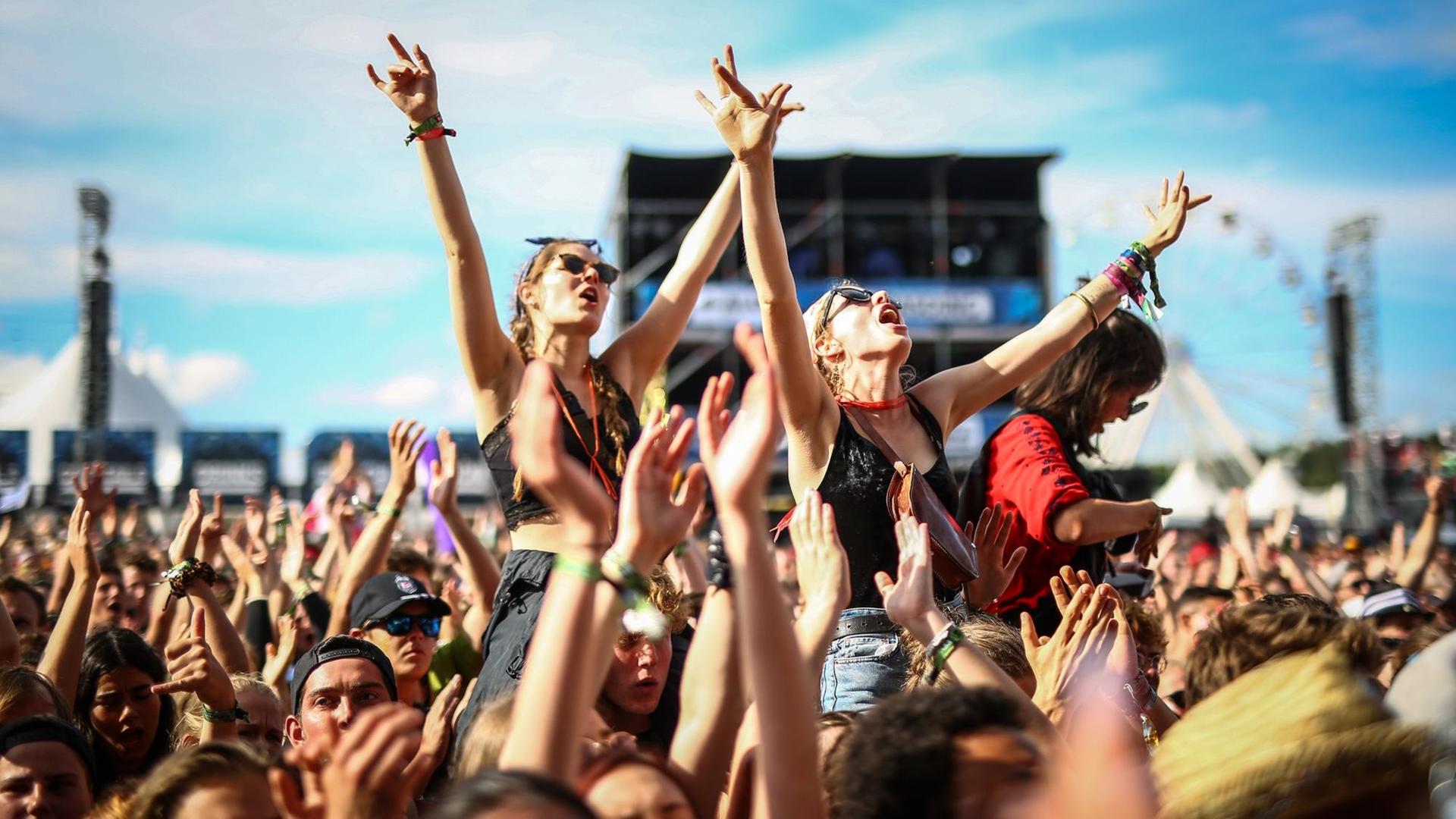 Das Bild zeigt die Menge des Southside Festival mit den Armen in die Luft gestreckt. Zwei Frauen stechen dabei besonders heraus: sie sitzen mit ausgebreiteten Armen auf den Schultern von zwei anderen Personen und singen zur Musik des Konzerts mit.
