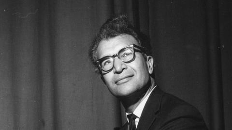 ein schwarzweißes Foto zeigt in seitlicher Porträtansicht einen Mann mit dicker Hornbrille in Anzug, Hemd und Krawatte. Er lächelt leicht.